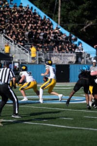 Football game action with two players in yellow uniforms, crowd and marching band in background.
