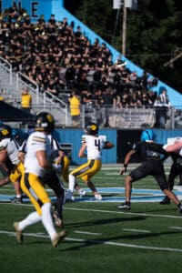 Football game action with player in yellow uniform ready to throw, band and audience in the background.
