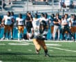 High school football player runs with ball on field during game, teammates and spectators in the background.