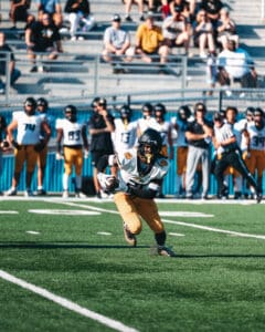 High school football player runs with ball on field during game, teammates and spectators in the background.