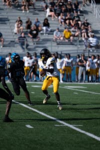 Football player in yellow and white uniform runs with the ball on a field, opposing team in pursuit.