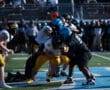 High school football game action, players in blue and black tackling opponents in yellow uniforms on the field.