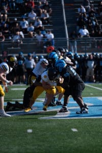 High school football game action, players in blue and black tackling opponents in yellow uniforms on the field.