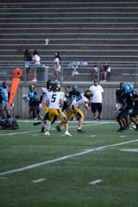 High school football game action with players in yellow and black uniforms running on field.