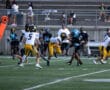 High school football game action, players in yellow and black compete on the field, bleachers visible in the background.