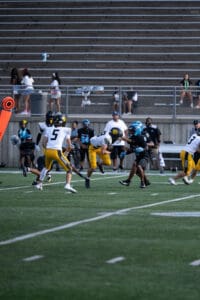 High school football game action, players in yellow and black compete on the field, bleachers visible in the background.