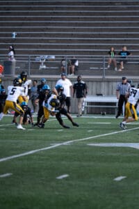 High school football game with players tackling on the field, empty bleachers in the background.