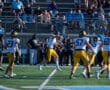 Football players in action on the field, with a crowd watching from the stands during a sunny day.