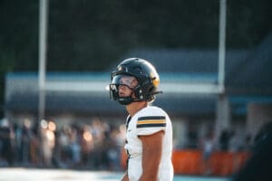 Football player on field wearing helmet and jersey in bright sunlight.