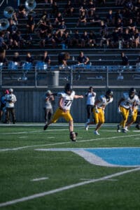 Football player kicking off in game, teammates running, band in stands watching.