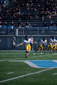 Football player kicking off a game on a sunny field, with a cheering crowd in the background.