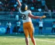 Football player in mid-air catches a pass during a game on a sunny day in a stadium with a crowd in the background.