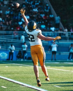 Football player in mid-air catches a pass during a game on a sunny day in a stadium with a crowd in the background.