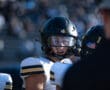 Football player in helmet and uniform listens intently during a game on a sunny day.