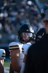 Football player in helmet and uniform listens intently during a game on a sunny day.