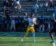 Quarterback in yellow pants throws a pass during a football game, with spectators and team on looking in the background.