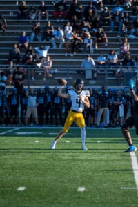 Quarterback in yellow pants throws a pass during a football game, with spectators and team on looking in the background.