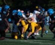 Football game action with players in black and blue uniforms tackling a player in yellow and white, holding the ball.