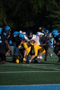 Football game action with players in black and blue uniforms tackling a player in yellow and white, holding the ball.