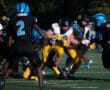High school football game action with players in yellow and black jerseys running through defenders in blue and black.