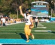 Football player celebrates touchdown in the end zone at outdoor stadium, sunny day game.