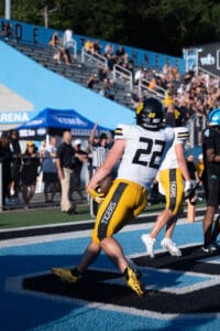 Football player in yellow and black scores a touchdown, pointing to the sky in celebration.