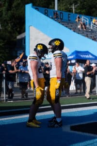 Football players celebrate on the field in matching uniforms and helmets, with a crowd and bleachers in the background.