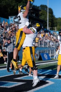 Football players in yellow celebrate a touchdown on the field with a jump and cheer in front of a cheering crowd.
