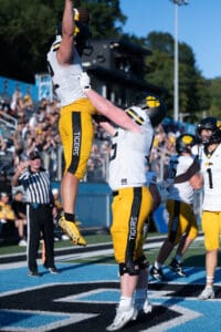 Football players celebrate touchdown with a lift during a sunny game day, fans cheering in the background.
