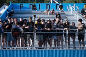 Enthusiastic crowd supporting a football team on bleachers, holding a helmet sign, wearing themed outfits.