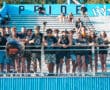 Group of enthusiastic sports fans in camo at a stadium, cheering behind a fence with blue bleachers in the background.