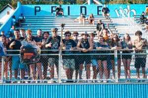 Group of enthusiastic sports fans in camo at a stadium, cheering behind a fence with blue bleachers in the background.