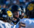 Football player in black helmet huddles with teammates, wearing uniforms with yellow paw print logo.