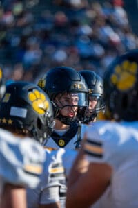 Football player in black helmet huddles with teammates, wearing uniforms with yellow paw print logo.