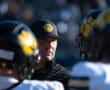 Football coach strategizing with players in helmets featuring yellow paw logos during a game on sunny day.