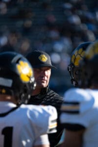 Football coach strategizing with players in helmets featuring yellow paw logos during a game on sunny day.