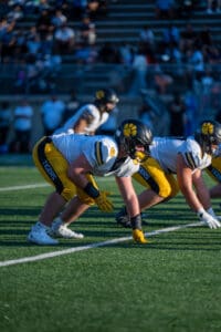 Football players in a ready position on the field during a game, wearing yellow pants and helmets with paw logos.