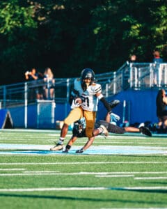 Football player in black and yellow evades tackle on field during a game, with spectators in background.