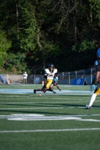Football player in white jersey runs downfield avoiding a tackle on a sunny day, surrounded by lush green trees.