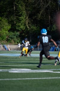 Football player in yellow pants runs with the ball on the field, pursued by opponents in blue helmets.
