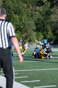 High school football game action with players tackling and referee observing on field.