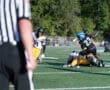 Referee watches as football players tackle during a game on a sunny day.
