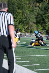 Referee watches as football players tackle during a game on a sunny day.