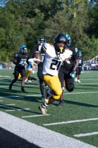 Football player in yellow pants running with the ball during a game, pursued by defenders in black uniforms.