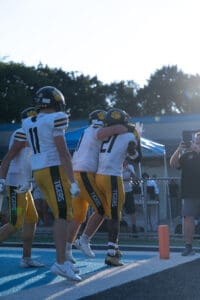 Football players celebrate a touchdown in the end zone during a sunny game, with a fan capturing the moment.