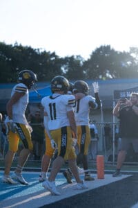 Football players in white and yellow uniforms celebrating a touchdown on the field, with a photographer nearby.