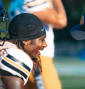 Football player smiling during a game, wearing a black head covering and team uniform.