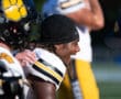 Football player smiling on sideline, wearing black helmet and gold uniform with paw logo, during a sunny game.