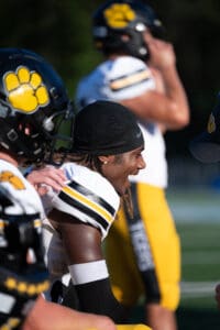 Football player smiling on sideline, wearing black helmet and gold uniform with paw logo, during a sunny game.