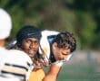 Football players in uniform, one smiling in foreground during practice, green field in background.